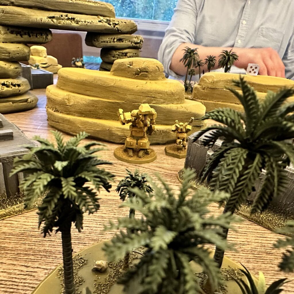 Heavy Gear Blitz wargame in progress. Shot is from amid the tabletop terrain, showing cliffs, palm trees, and buildings, between which a Heavy Gear patrol is advancing.