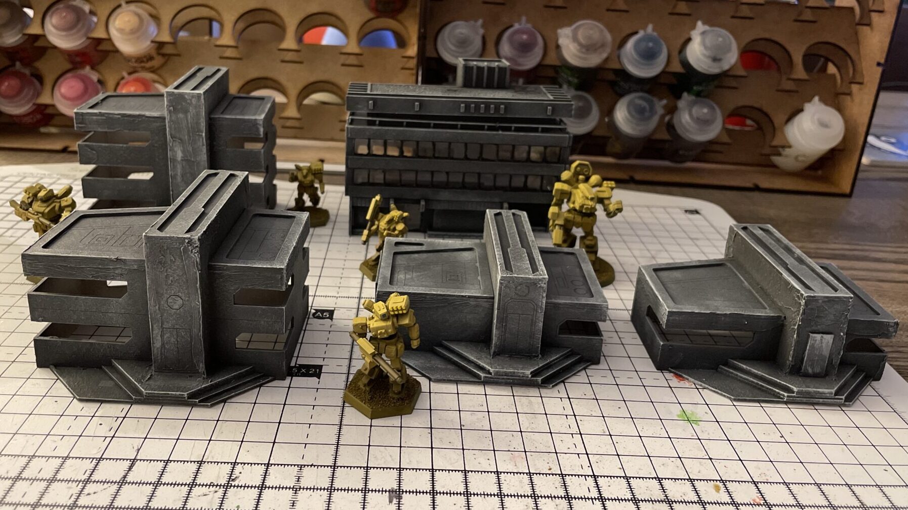 Work in progress 12 mm scale wargaming terrain, with Heavy Gear miniatures for scale. Five buildings of different makes, all painted a similar gray.